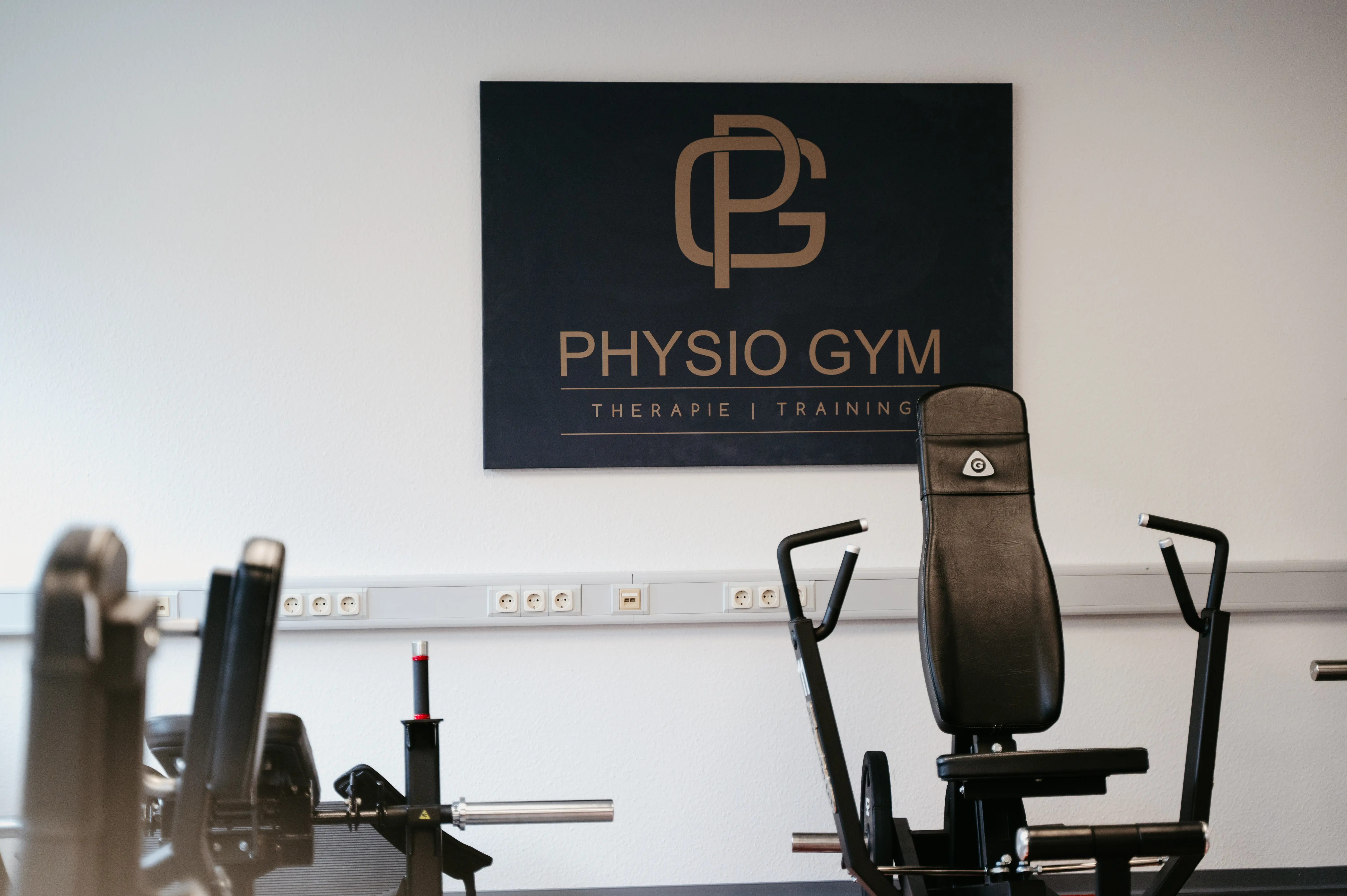 Bild des PhysioGym Logos hängt an einer Wand. Im Vordergrund stehen Traingsgeräte.